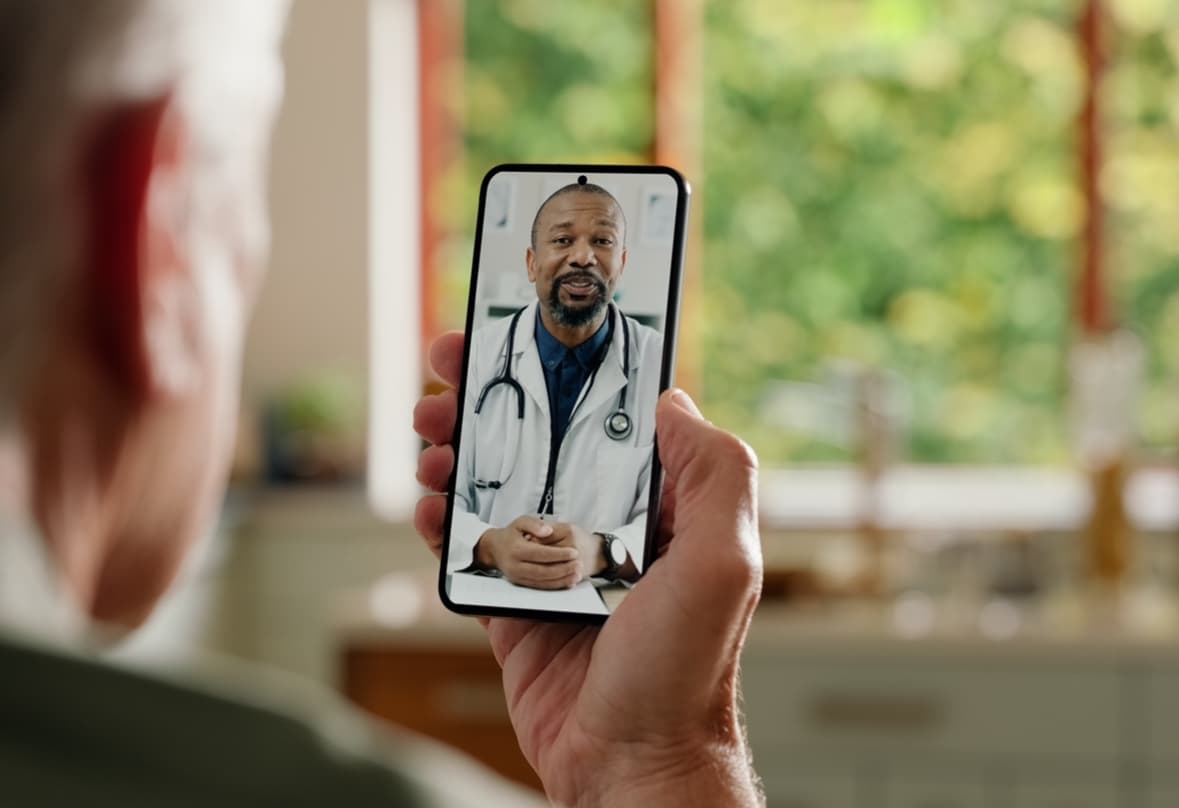 Elderly person holding a smartphone during a virtual consultation with a male doctor in a white coat, representing telehealth and remote medical care.