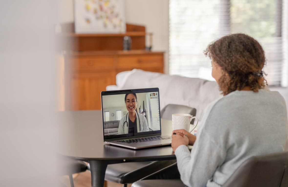 Woman sitting at a table at home, having a virtual consultation with a smiling female doctor on a laptop screen, representing telemedicine and remote healthcare services.