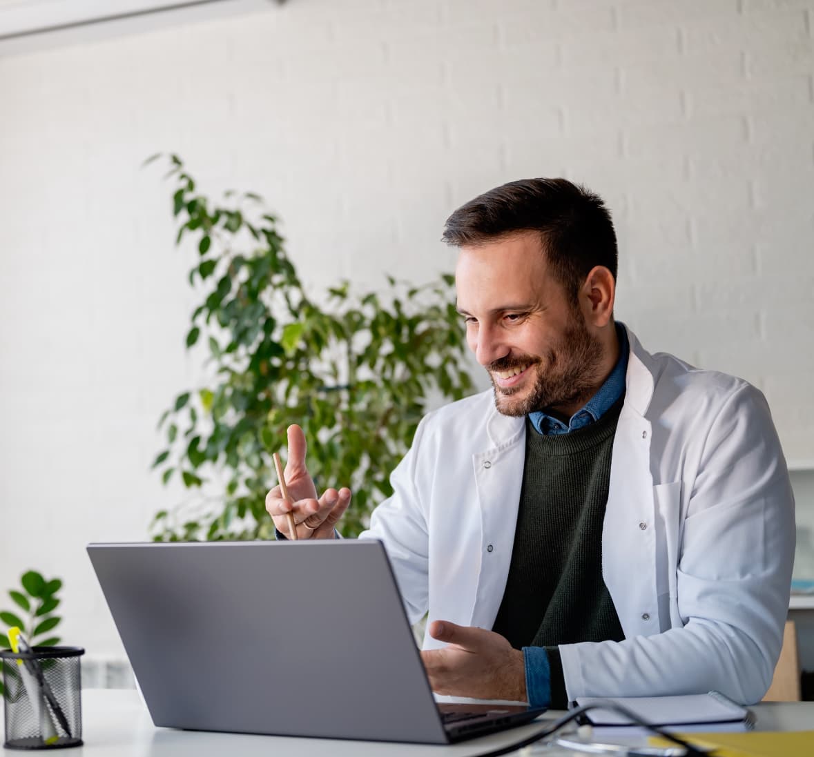 Smiling doctor wearing a white coat conducting a virtual consultation on a laptop, sitting in a modern office with plants in the background, representing telemedicine and online healthcare services.